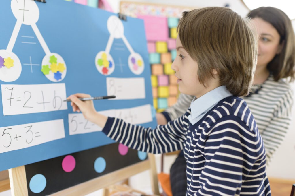 Cute boy solving graphic math exercises at kindergarten, under educator observation